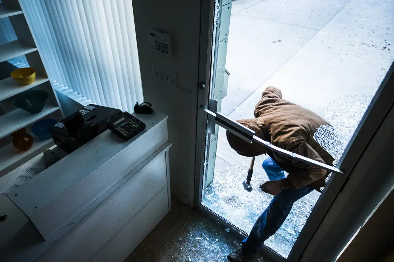 A burglar in a brown hoodie breaks into a store by smashing a glass door with a crowbar.