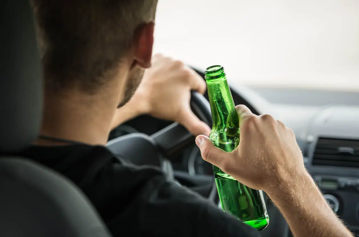 A man drives a car while holding a green bottle, drinking and driving. Suggesting he is driving under the influence of alcohol