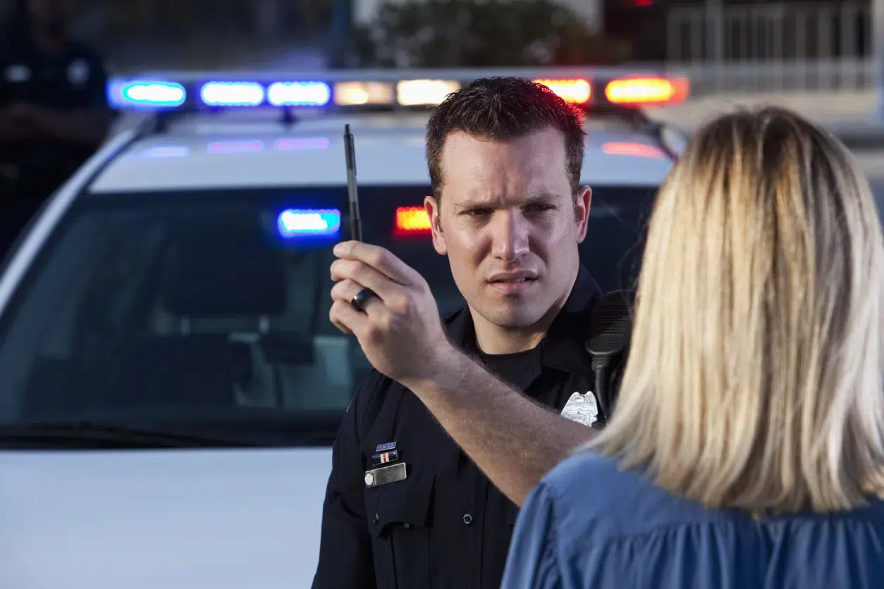 A police officer conducts a field sobriety test with a pen in front of a woman. A patrol car with flashing lights is visible in the background.
