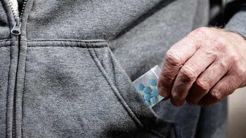 In a case illustrating drug crimes, a hand slips a small bag of blue pills into the pocket of a gray hoodie.