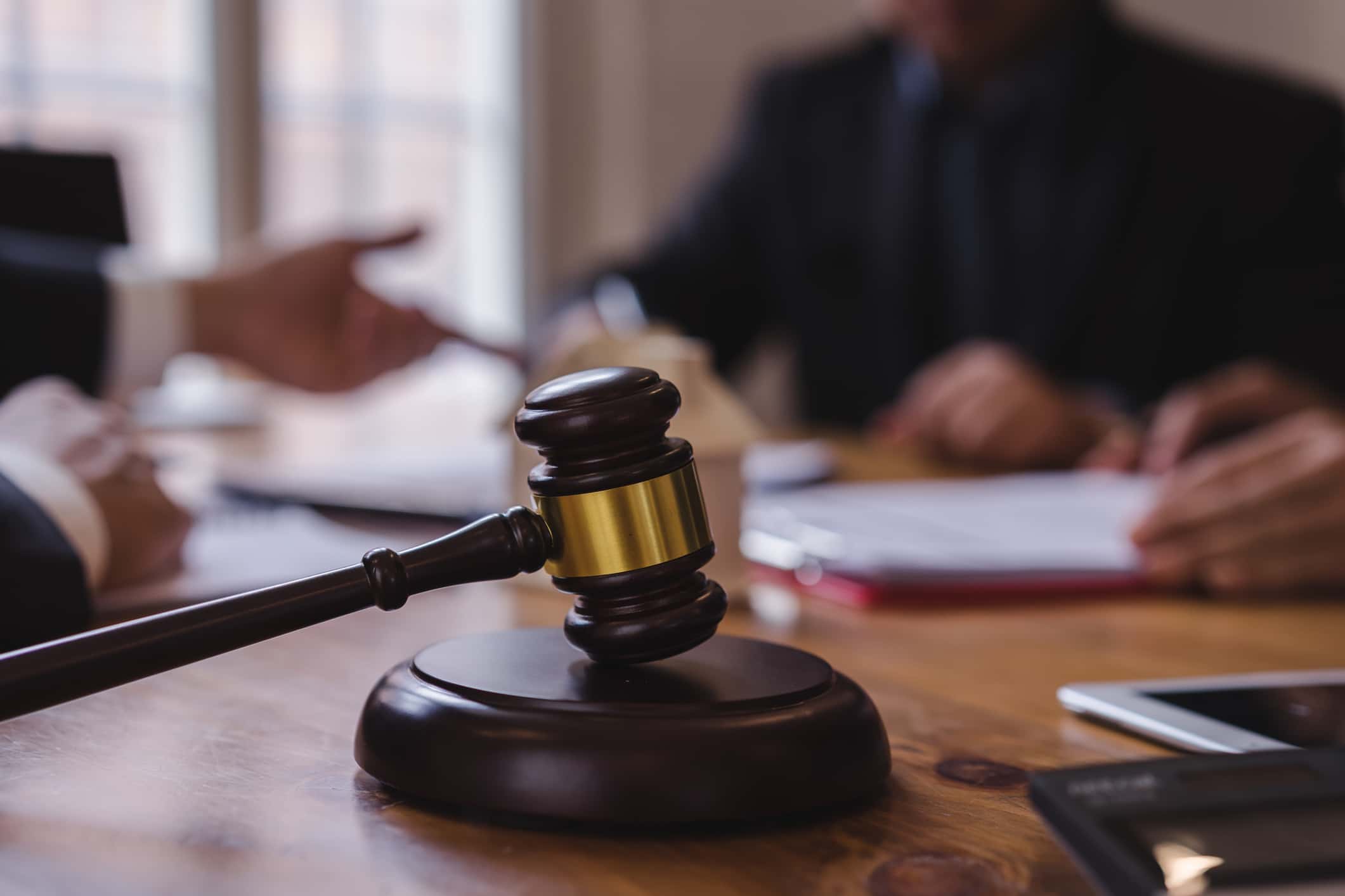 A judge's gavel rests on a wooden table in a courtroom setting. In the background, blurred criminal defense lawyers in suits discuss