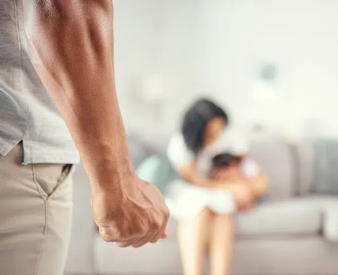 A tense domestic violence scene shows a close up of a clenched fist in the foreground, while a woman holding a child sits on a couch in the blurred background.